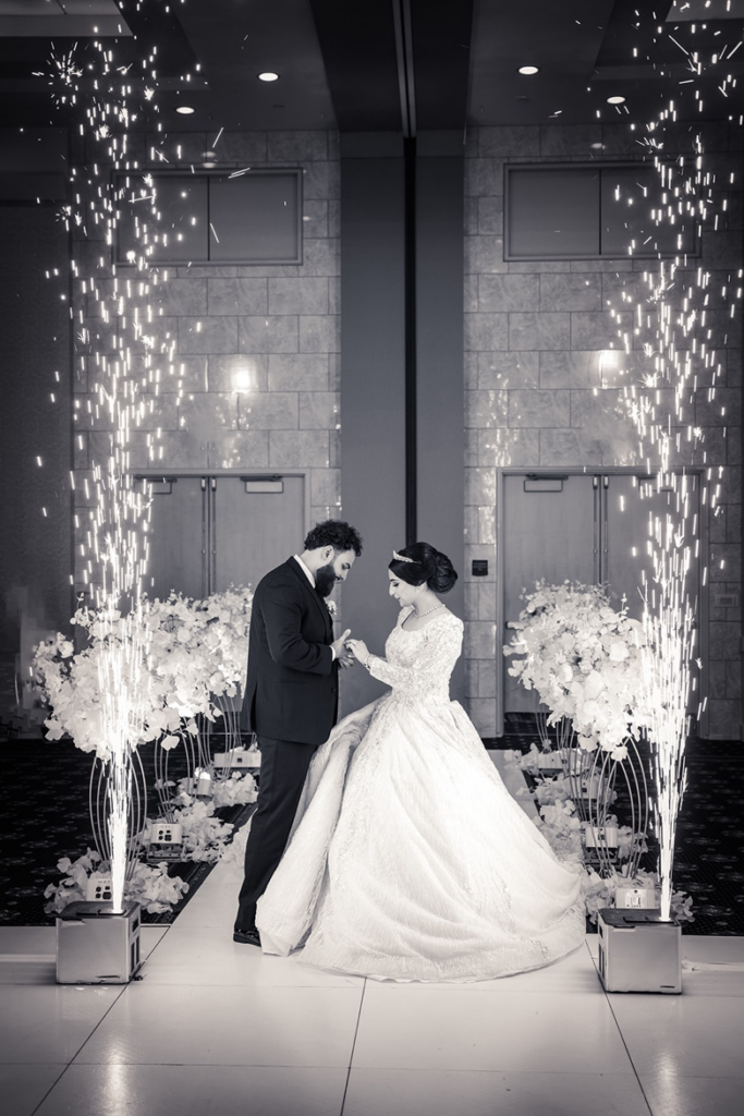 Black and white photograph of a bride and groom standing on a custom dance floor wrapped in white while indoor cold spark fireworks shoot up beside them.